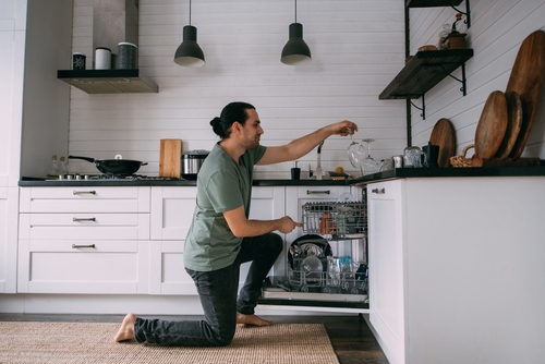 Young brunette Caucasian man unstacking plates from the dishwasher whilst kneeling on the kitchen floor