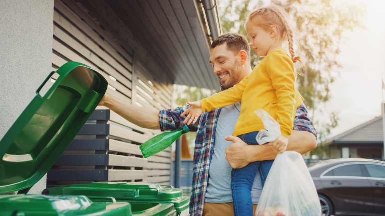 Caucasian father holding his young daughter whilst throwing away an empty bottle and food waste into the correct recycling bin outside their house