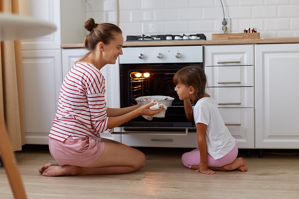 Caucasian mother and her young daughter kneeling beside the gas oven in their kitchen looking at pastry they baked together